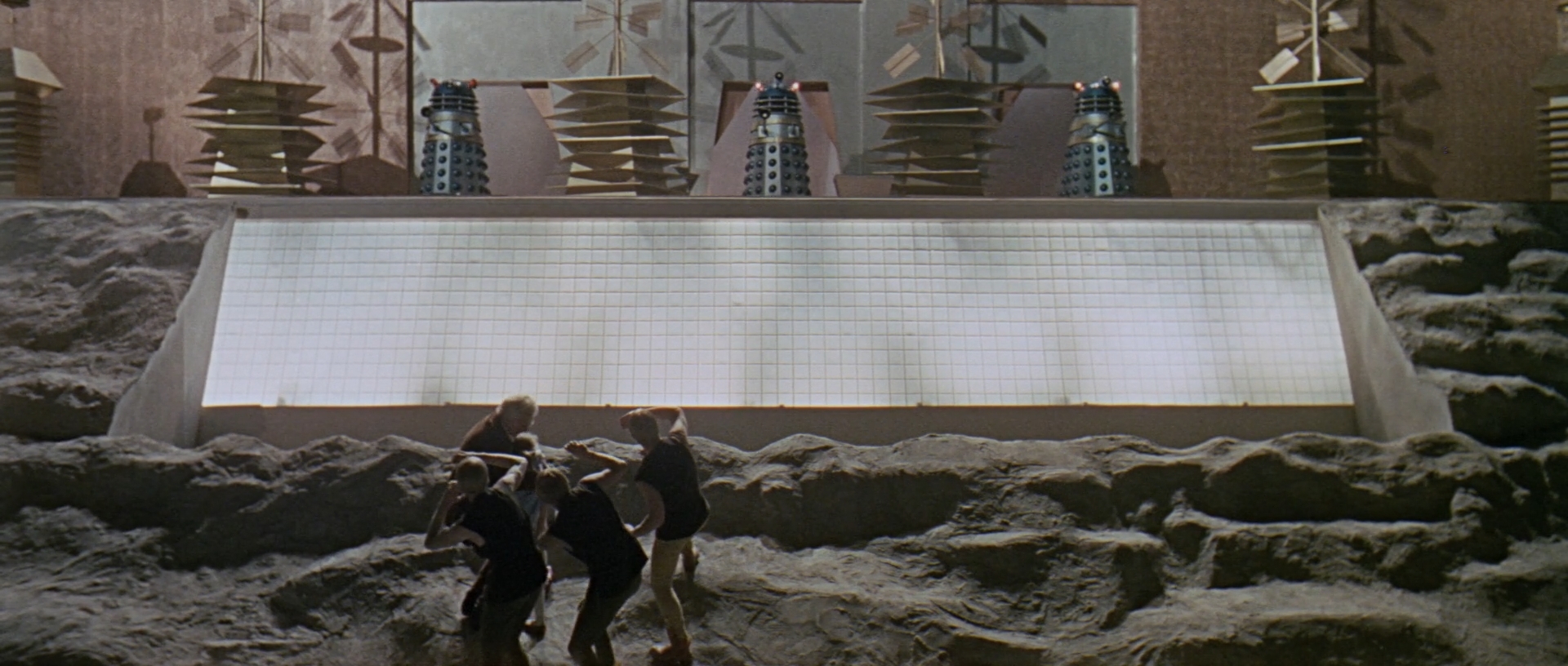 Image description: Exterior of the Dalek city. Three Daleks emerge from three doors on a raised platform. Below them, there are bright lights, and four people shield their eyes from them below.