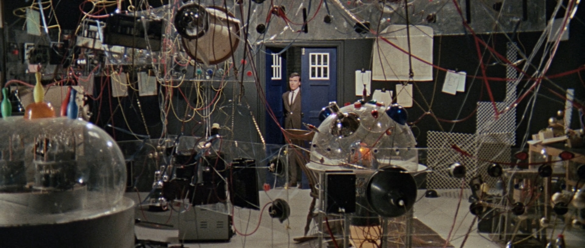 Image description: In the foreground there is a lot of scientific equipment and wires dangling from the ceiling. Ian looks into the room through the door of Tardis in the background.