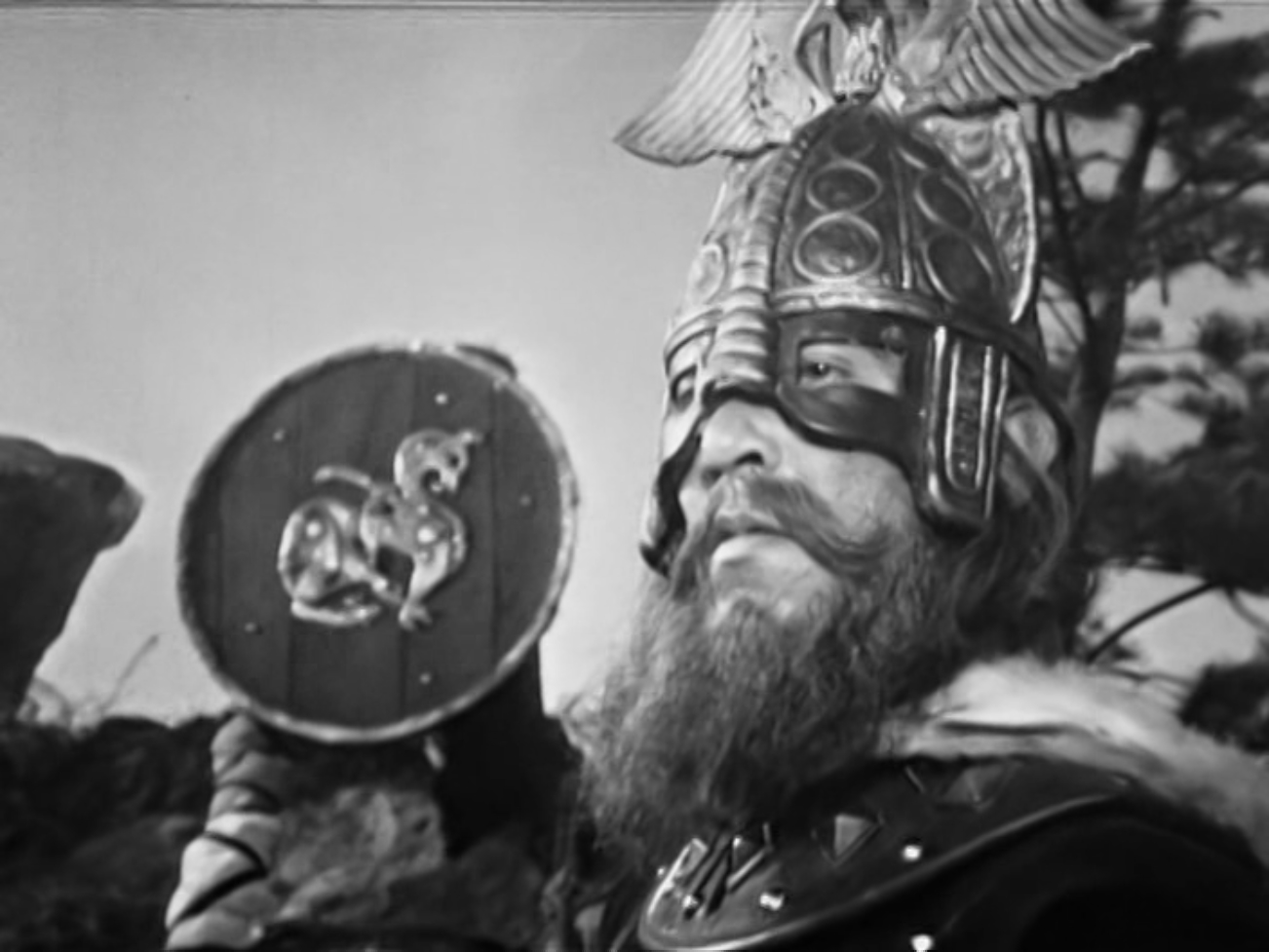 Image description: The Viking leader wearing an elaborate helmet topped with an eagle. In the background, another Viking is hidden behind a decorated shield.