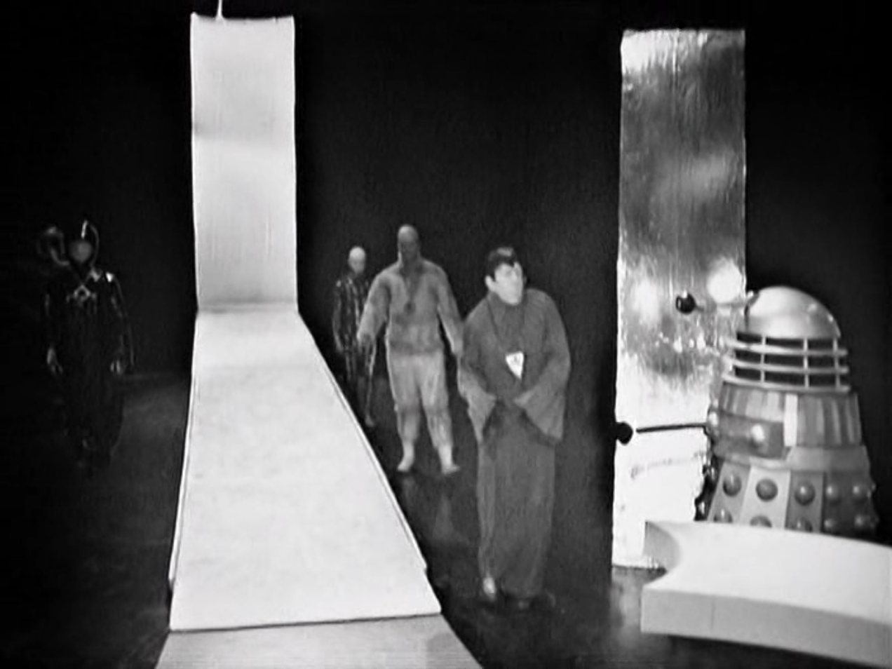 4 humanoid aliens approach a table with a Dalek waiting to greet them.