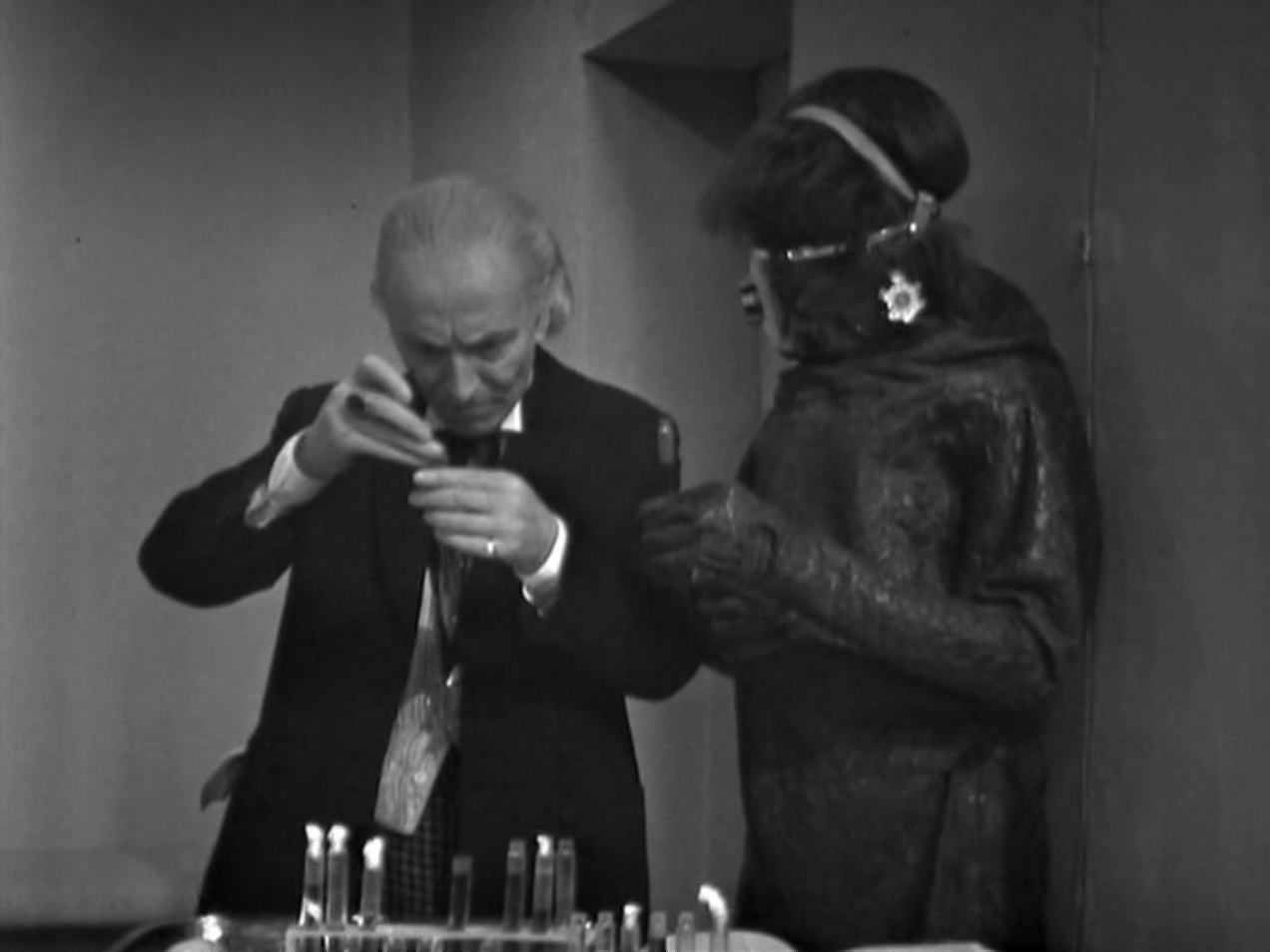 The Doctor adds an ingredient to a test tube as a Monoid looks on.