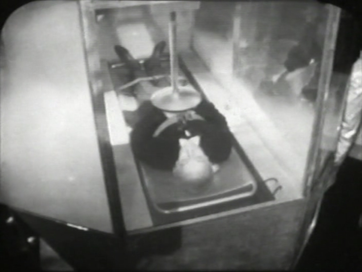 The Doctor lies strapped to a table within a glass case. A machine is positioned above his chest.