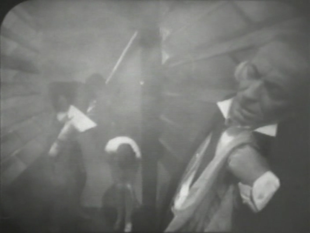 A smoky corridor. The Doctor is slumped against the wall in the foreground, and Steven and Dodo are in the background. Steven has a gun, and Dodo is bent double.