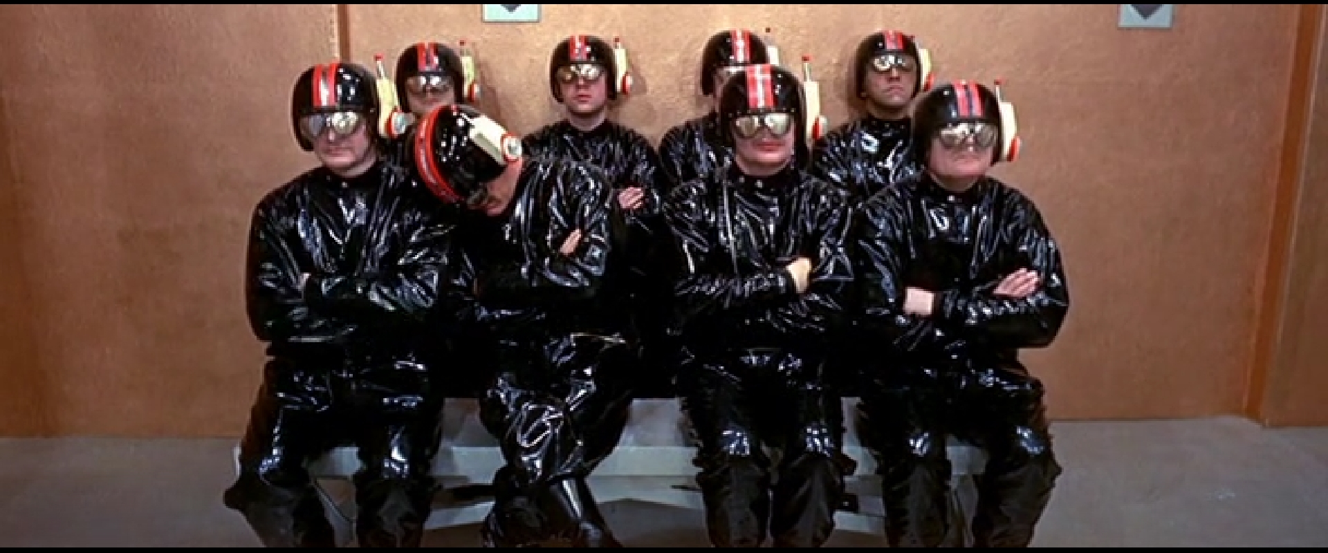 8 men in black latex overalls and helmets with visors sit on two rows of benches with their arms folded. Tom (front row, second from left) is asleep, leaning on his neighbour's shoulder.