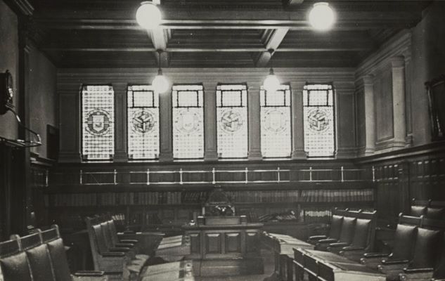 Photo of an old fashioned room with stained glass windows and a large number of chairs. This is the chamber of the House of Keys, Manx’s Lower House