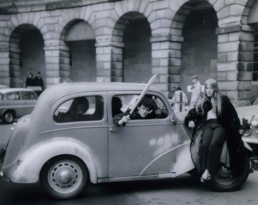 A small car in front of a Georgian terrace building. The driver is waving a large sword. 