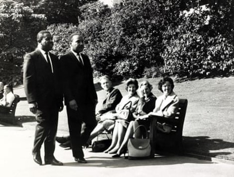 Martin Luther King in London 1964 walking through a park whilst onlookers watch from a bench.