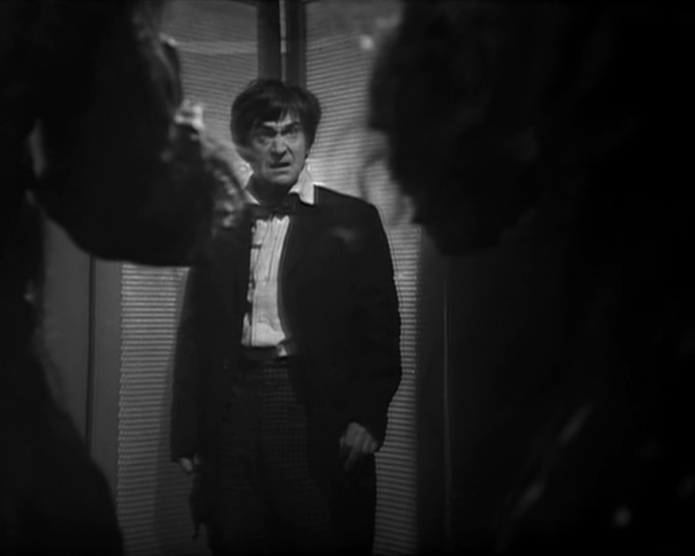 The Doctor stands with his back to a wall, looking apprehensive. In the foreground, obscured, are a pair of Ice Warriors at either side of the shot, framing him. 