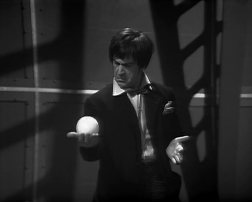 The Doctor stands alone, examining a small white sphere in the palm of his hand.