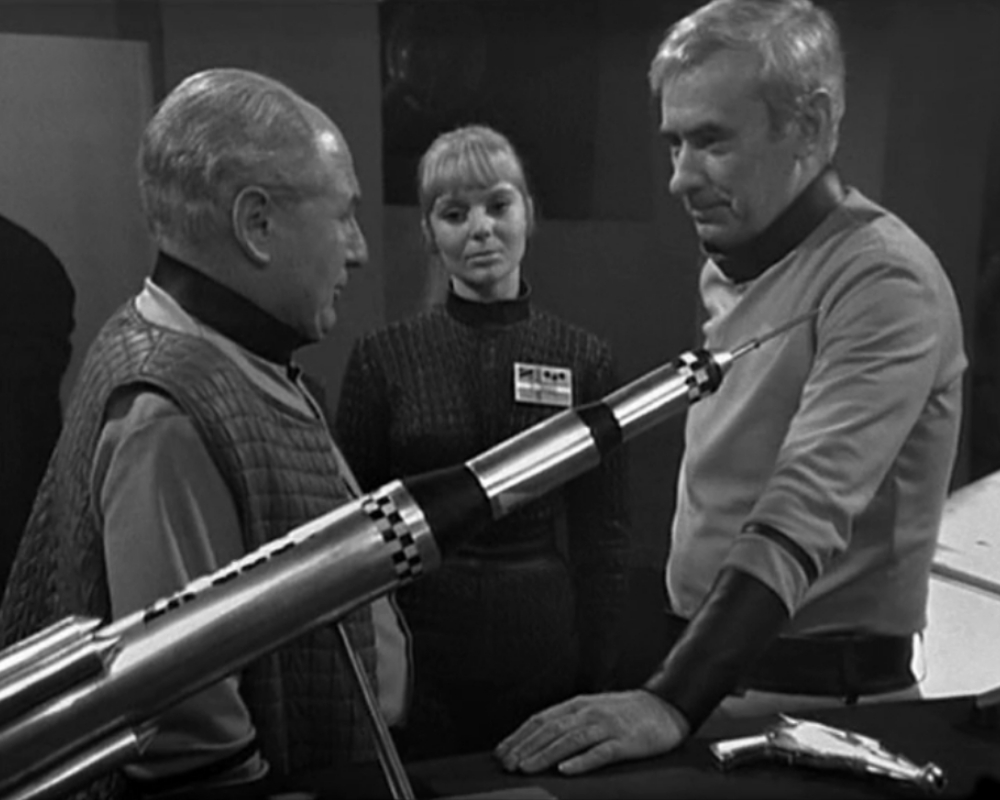 ID: Professor Eldred speaks to Commander Radnor. Kelly stands between them. There is a model rocket in the foreground. 