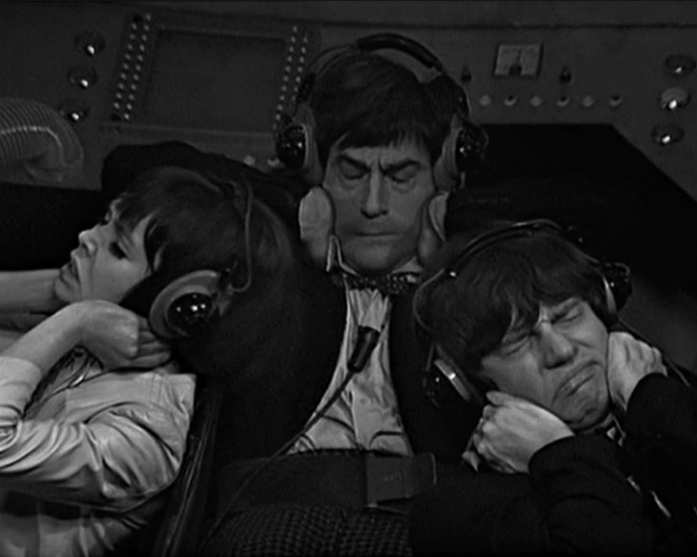 ID: Left-Right: Zoe (late teens, dark hair, white), the Doctor, Jamie (early 20s, dark hair, white). All three are seated in a small cockpit. All three are wearing headphones. All three have looks of discomfort on their faces.