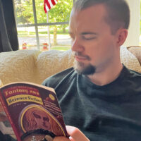 A white man with dark short hair and a dark van dyke beard sits on a yellow couch reading a fantasy periodical.  A window in the background shows an empty suburban street.