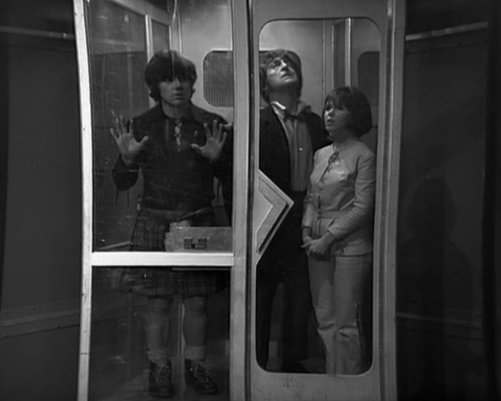 ID: Jamie (white male, dark hair, young adult), the Doctor (white male, dark hair, middle-aged) and Zoe (white female, dark hair, young adult) stand in a glass box, similar to a phone booth.