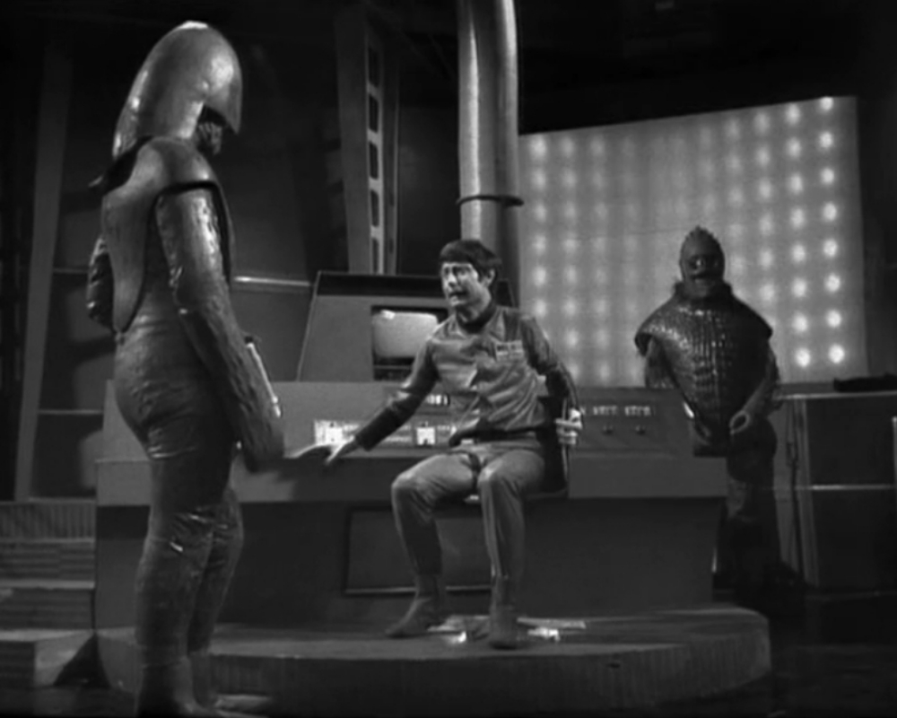 ID: Fewsham (white male, 30s) sits in front of a computer terminal between two Ice Warriors (left foreground, right background, both wearing scaly armour and helmets)