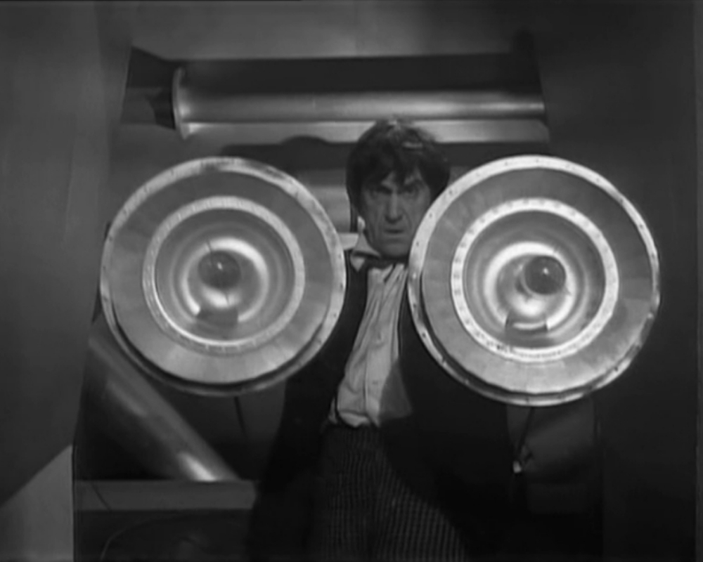 The Doctor holds up two round metal dishes with lightbulbs in the middle.