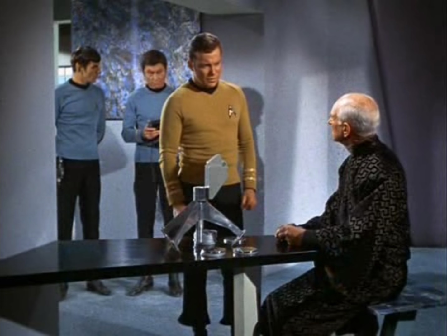 Spock and McCoy stand behind Kirk, who is looking down at Mr. Atoz, a balding, white-haired man in a black gown, sitting at a table with some kind of viewer and mirror-surfaced disks