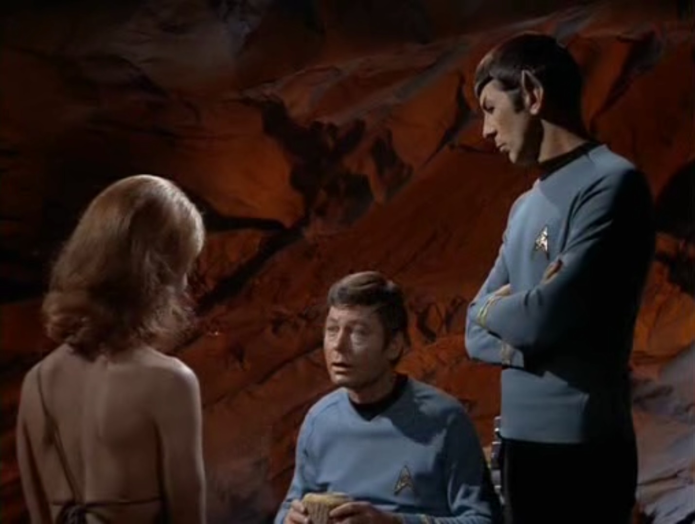 A seated McCoy talks to Zarabeth, viewed from behind, wearing a fur bikini, a Spock looks at him with folded arms, in a red-lit cave