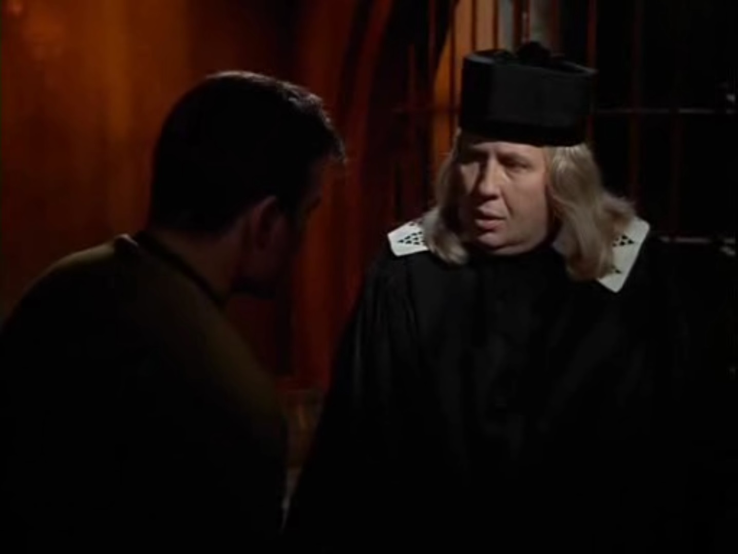 A florid, long-blond-haired, older man in a black hat and robe visits Kirk in jail