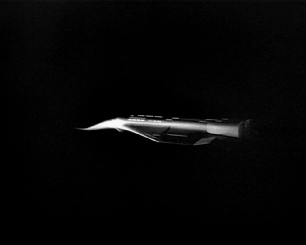 ID: Monochrome photo, sleek dark spaceship against a black void. The ship resembles a jet plane with an angled nosecone.