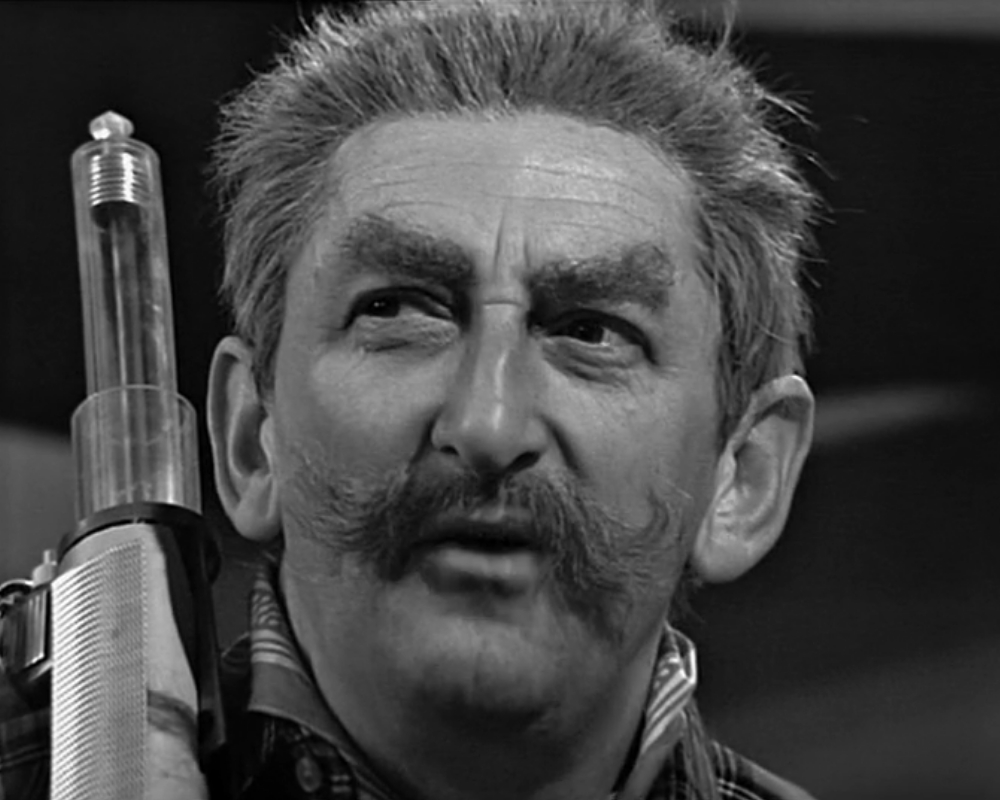 ID: Monochrome photo, close-up of Milo Clancey (Gorden Gostelow) looking off to his side with his eyes narrowed in suspicion. He has a futuristic gun with a plastic barrel on his shoulder. Clancey is a white man in his late 50s-early 60s, with short bristly hair, large eyebrows and an impressive moustache with the ends curled up. He is wearing a checked shirt.