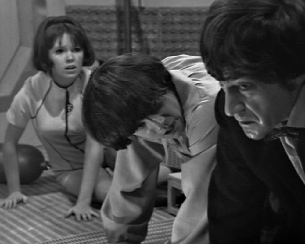 ID: Monochrome photo, Zoe (young white female, dark hair), Jamie (young white male, dark hair) and the Doctor (middle-aged white male, dark hair), all on hands and knees, all appearing distressed.