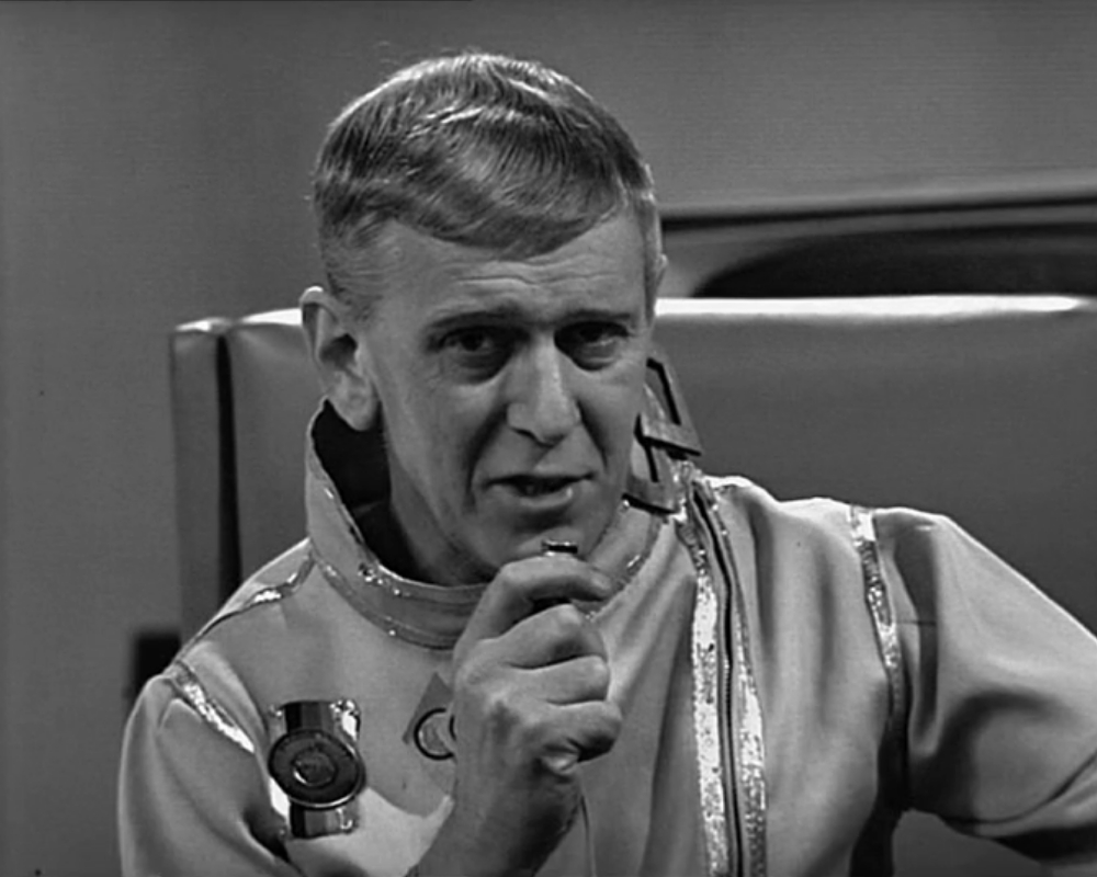 ID: Monochrome photo, General Hermack (Jack May), speaking into a receiver. He has neat hair greying at the temples, and wears a high-collared spacesuit-like garment with metallic trim. He is white and appears to be in his fifties.