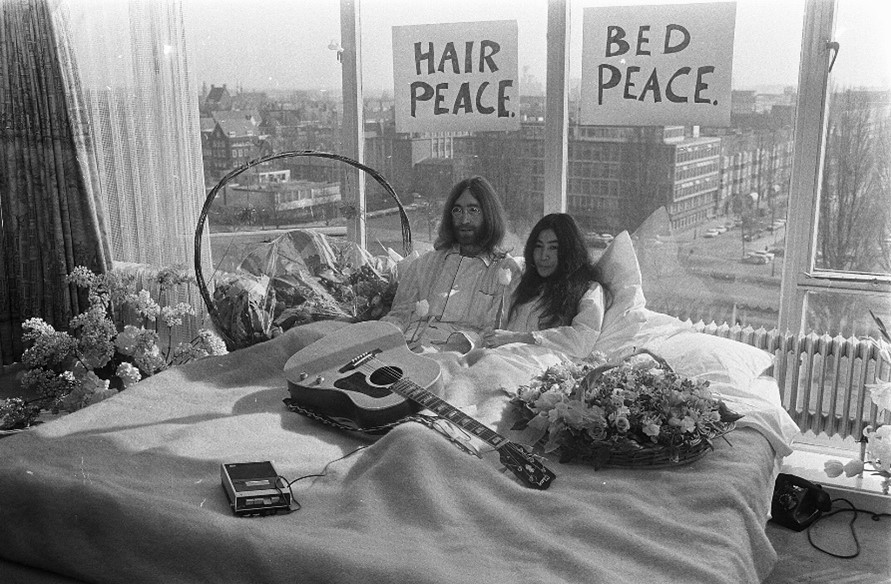 Yoko Ono and John Lennon at the Amsterdam Hilton in pyjamas in bed with signs pasted on the window 
saying "hair peace" and "bed peace"