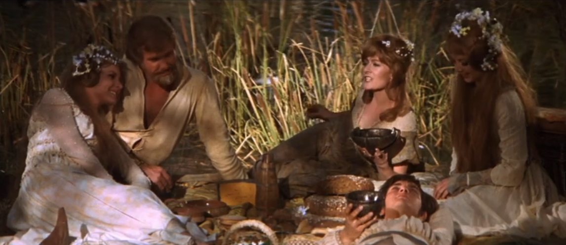 An image from the movie "Camelot" showing several maids and young men adorned with flowers, lounging around outside and having a picnic in the grass.