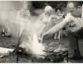 Black and white photo of people sitting in plastic chairs around a grill with various meats on it