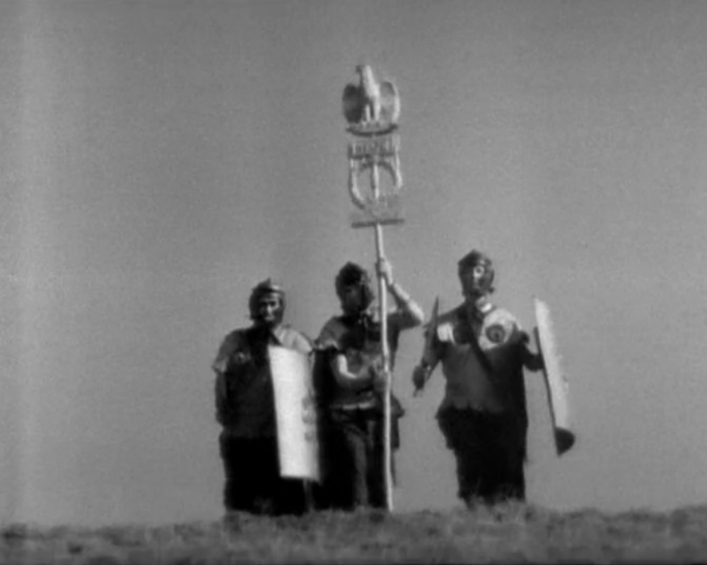 Three Roman soldiers bearing an eagle standard standing on the crest of a hill.