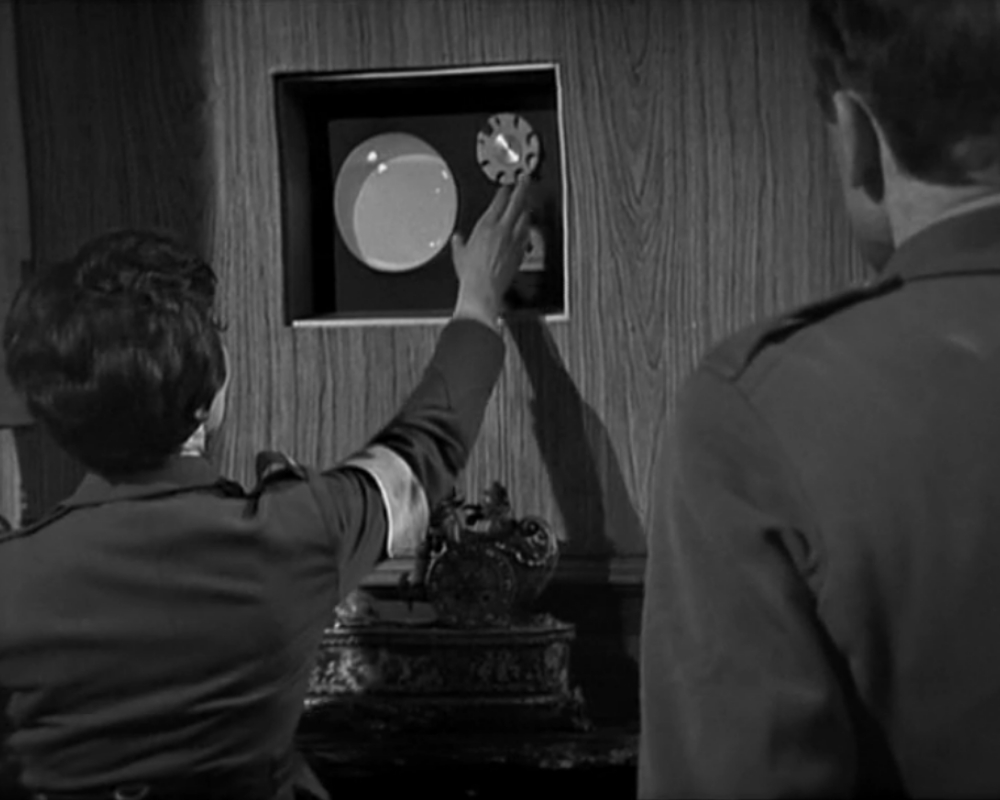 The General's Office. There is a round glass screen set into a wall panel. Lady Jennifer, her back to the camera, reaches up to touch it. Lt. Carstairs, seen from the back, is at the edge of the frame.