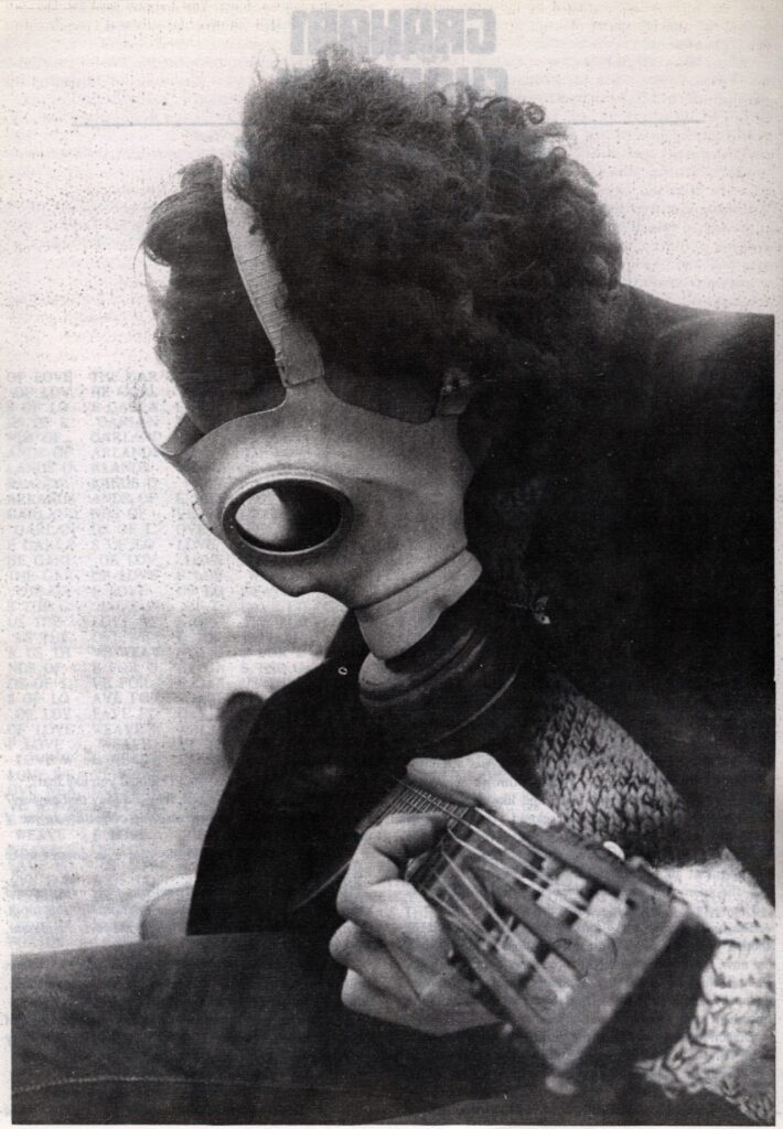 IMAGE: A photograph of a man with curly hair playing a guitar whose face is obscured by a gas mask. 