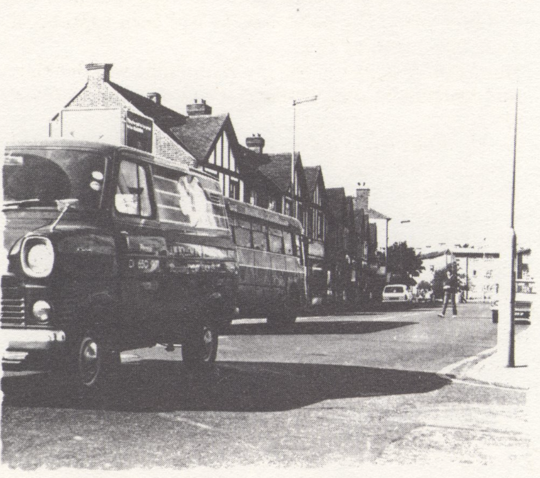 Photo by Roy Cornwall BW photograph of a street. There are houses and vehicles. A pedestrian is crossing the street in the background.