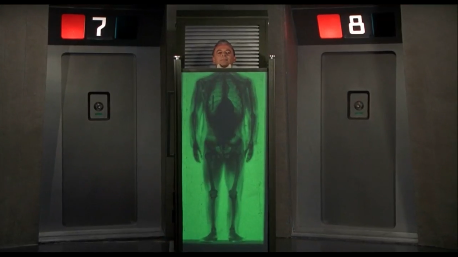 Photo of a scene. A man is standing behind an X-ray scanner, his head, chin up, visible above the machine. We see his squeleton through the green glass, a pen is visible on his chest, in front of his right lung.