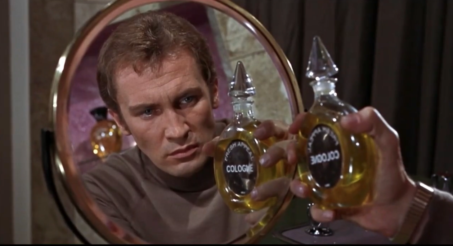 A photo of a scene. Ross is in front of a mirror. The label on the bottle of cologne he is holding is backwards and the reflection in the mirror shows the readable label.