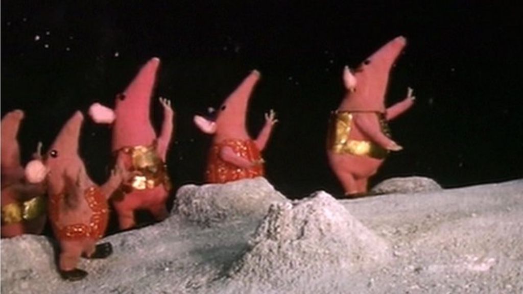 Photo from the show. Standing on a desert grey ground, pink mice-shaped aliens with red and gold vests are looking up and raising their arms. There are a few stars visible in the sly. The aliens seems to be made out of fabric.