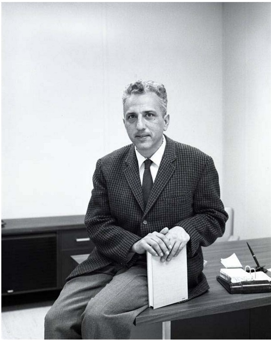 Dr. Edward D. Goldberg, a white man with gray hair in a suit and tie.  He is sitting on a desk, holding a book, and smiling.