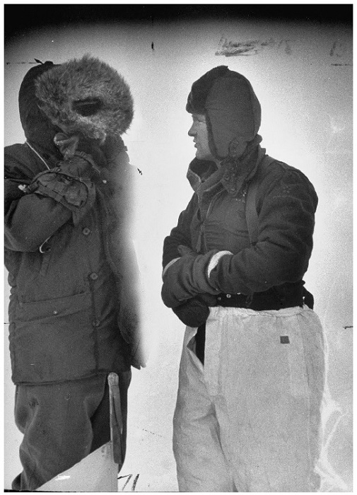 A photograph of Col. J. O. Fletcher, a white man  wearing snow pants, a thermal undershirt, suspenders, and a winter hat.  He is having a conversation with a second unidentified person who is completely obscured by their parka hood.