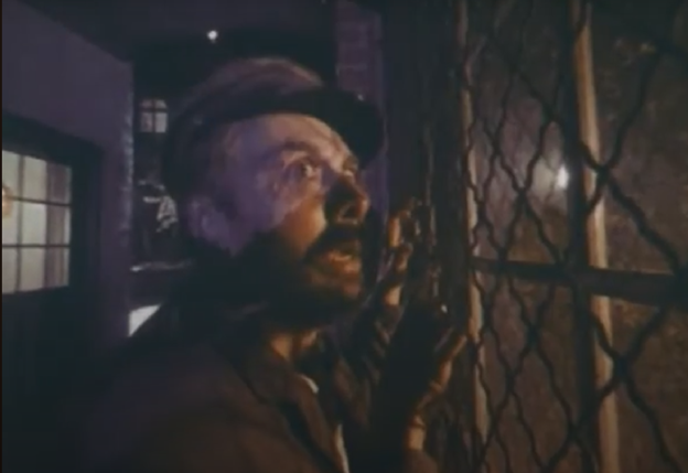 Still frame from the TV series Night Gallery. It shows a man standing outdoors at night, looking in anxiety at whatever could be on the other side of a wire mesh fence.