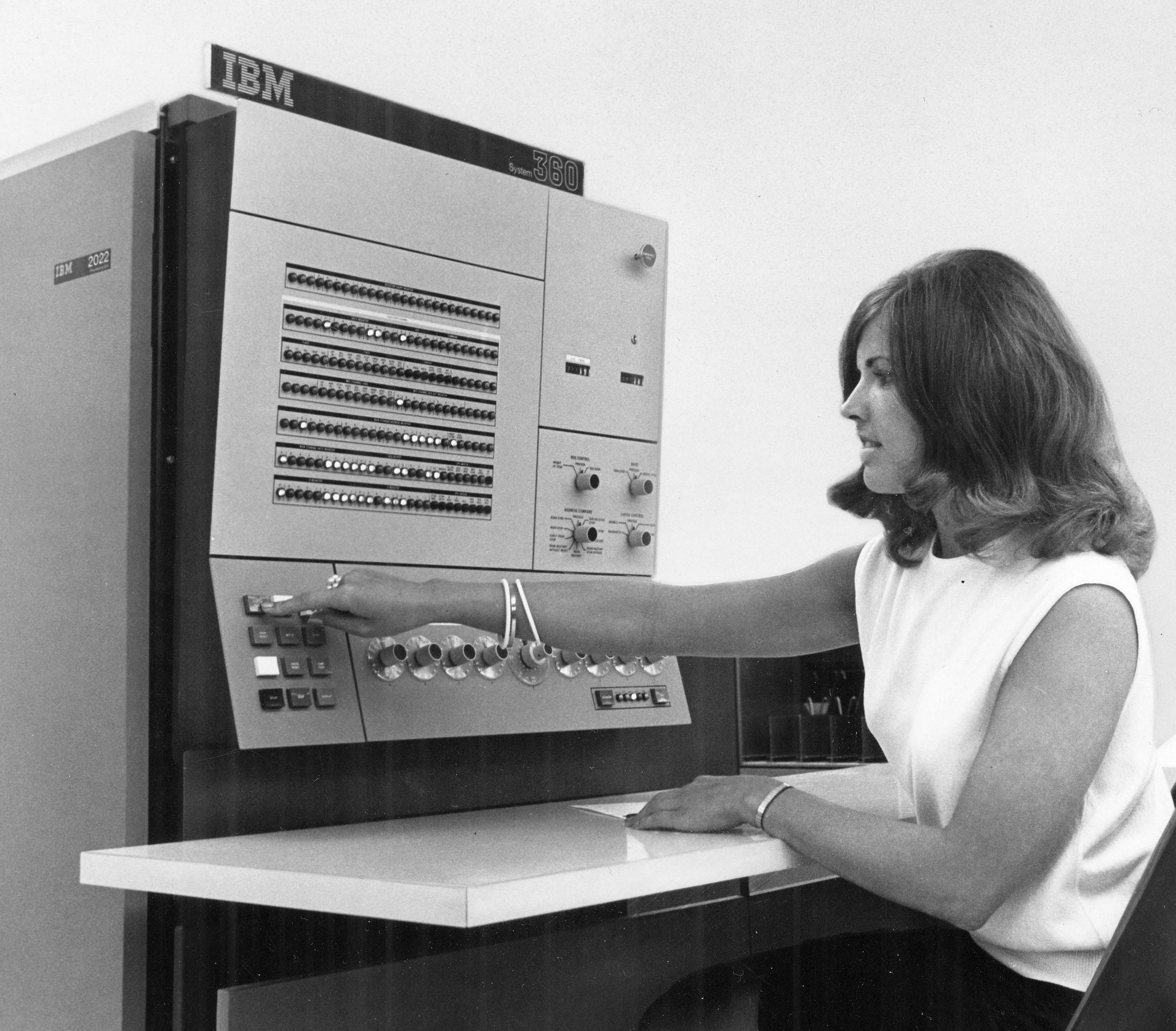 Black-and-white photograph of a young woman sitting at an office, working with an old computer that is the size of a refrigerator.