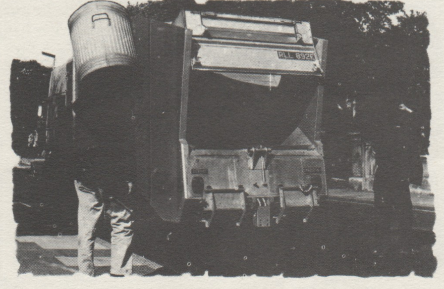 Black-and-white photograph of a man taking a garbage can to the back of a garbage truck.