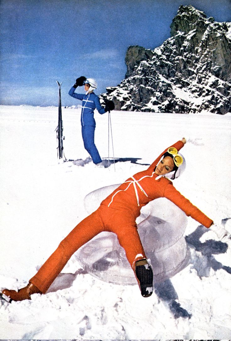 Photograph of two women skiing. They're on a snowy landscape, wearing two-piece jumpsuits (one woman wearing all blue, the other all orange) and protective helmets. The woman dressed in blue is standing in the background and looking into the distance. The woman dressed in orange is simulating a falling position with spread arms and legs. There is a tall rocky formation covered in snow farther in the background.