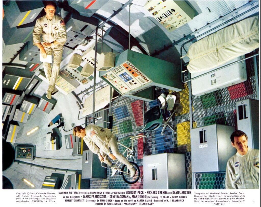 A promotional image of the movie. Three characters are seen in a huge room with a circular ground/wall. There is a technical console and a chair, camp beds. One of the characters is exercising on a stationary bike.