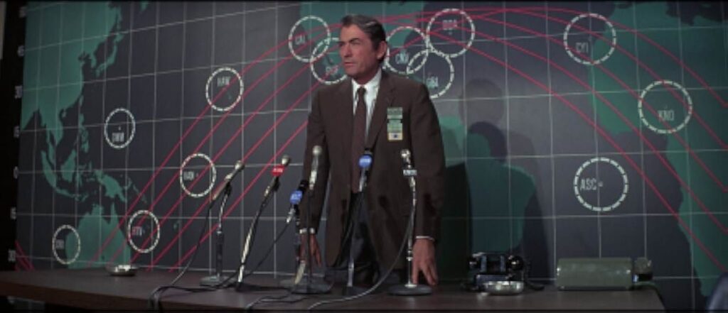 A character in a brown suit is standing in front of several microphones for what seems to be a press conference. The wall behind him consists of a world map with red parallel curbed lines. 