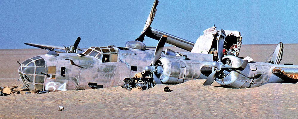 A color photograph of the wreck of the bomber 'Lady Be Good' as it lays on the sand in the Libyan desert. The back half of the plane lies at right angles to the front half, and there are several small items scattered around the main wreck.