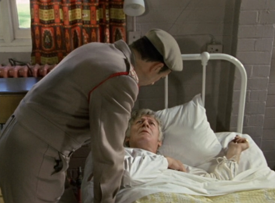 The Brigadier leans over the Doctor's hospital bed, as the Doctor frowns up at him.