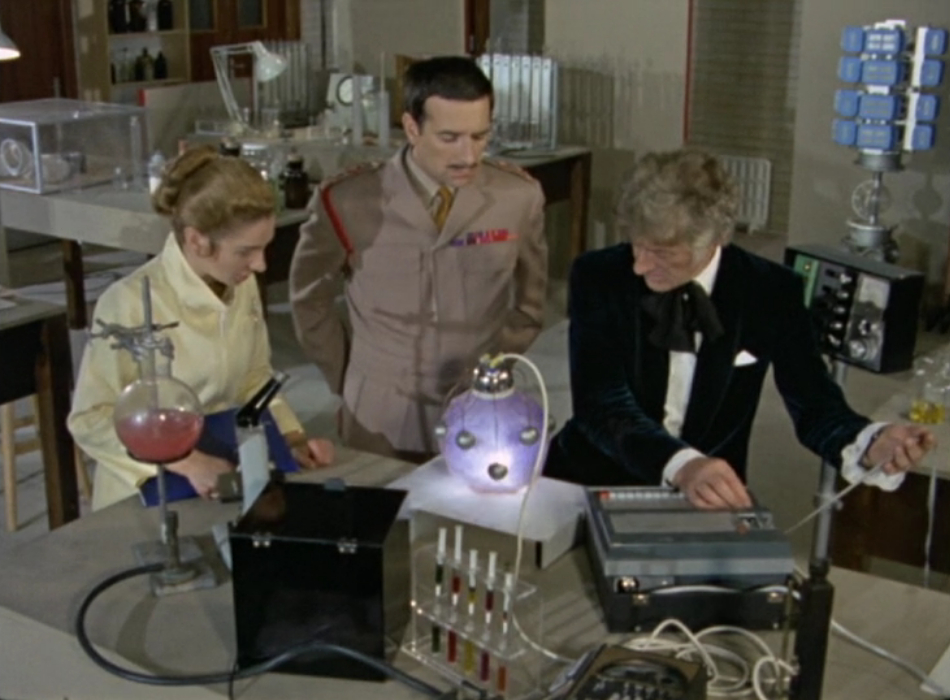 Left-Right: Liz, the Brigadier and the Doctor. They're looking at a glowing orb-shaped object which is connected to various scientific equipment.