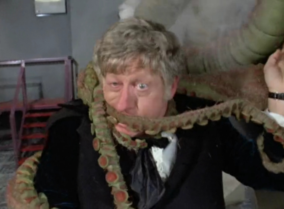 The Doctor being strangled by green tentacles, with a comic expression of distress on his face. 