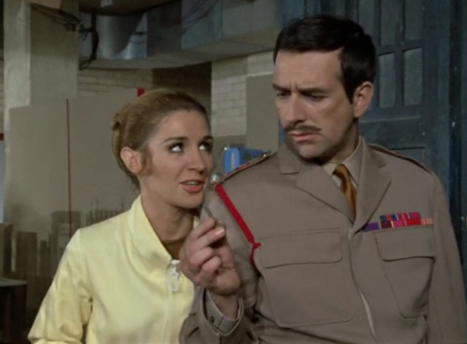 Liz Shaw (left) and the Brigadier (Right). The Brigadier is frowning at the TARDIS key in his hand, and Liz stands behind him with a slightly mocking look on her face.