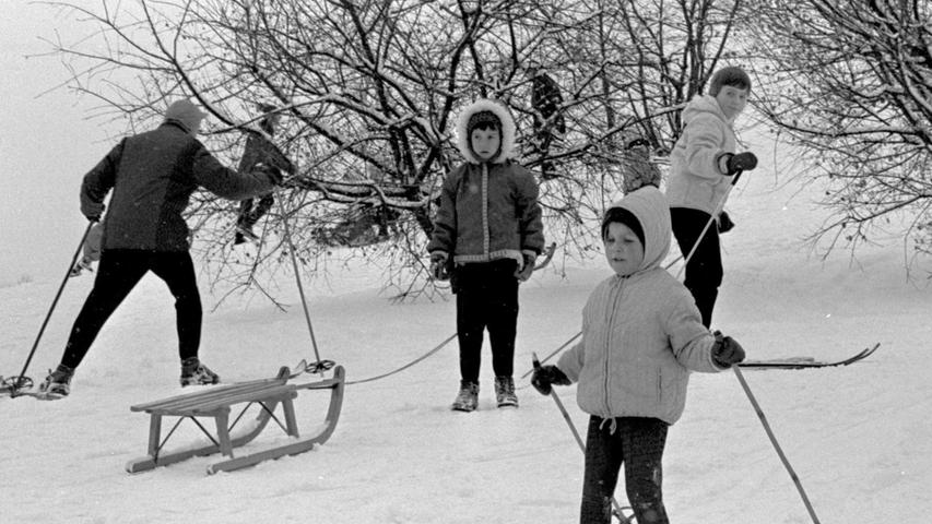 A child stands on skis, while another pulls a sled.